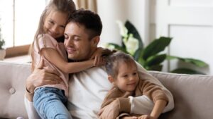 Padre abrazando a sus dos hijas en el sofá de su casa gracias a la crianza respetuosa